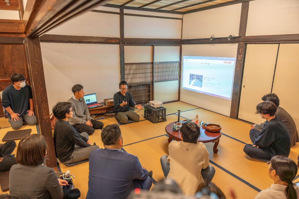 歴史を感じさせる木造の和室にて、壁面に投影されたウェブサイトの画面を囲むように参加者が座り、解説を行う男性の身振り手振りを交えた話を全員で見ている談会の様子の写真