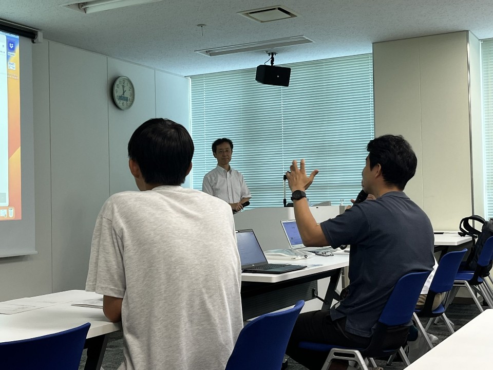 研修会場で前方に立つ男性に向かって、手前に座る参加者がノートパソコンを広げながら片手を挙げて質問や発言をしている様子の写真