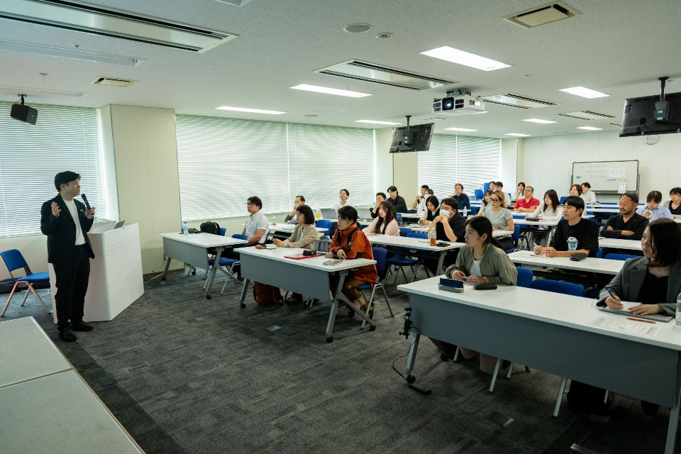 研修会場の前方でマイクを手に話す講師の男性と、デスクに座りながらメモを取ったり前方を見つめたりして熱心に講義に耳を傾けている多くの受講生たちの様子を広角で捉えたセミナー風景の写真
