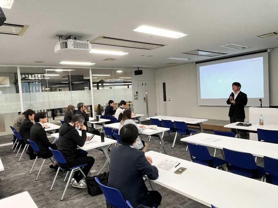 研修会場で、前方のスクリーンに映し出されたロゴを背にマイクを持って話す男性と、その話をデスクに座って真剣に聞き入るスーツ姿の参加者たちの様子を後方から写した写真