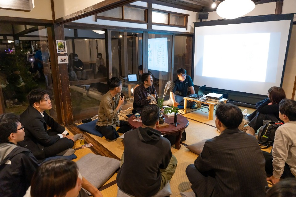 夜の落ち着いた和室にて、プロジェクターで投影された資料を見つめながら円卓を囲む参加者たちが、マイクで発言する人の話を熱心に聞き入る様子を捉えた座談会の様子の写真