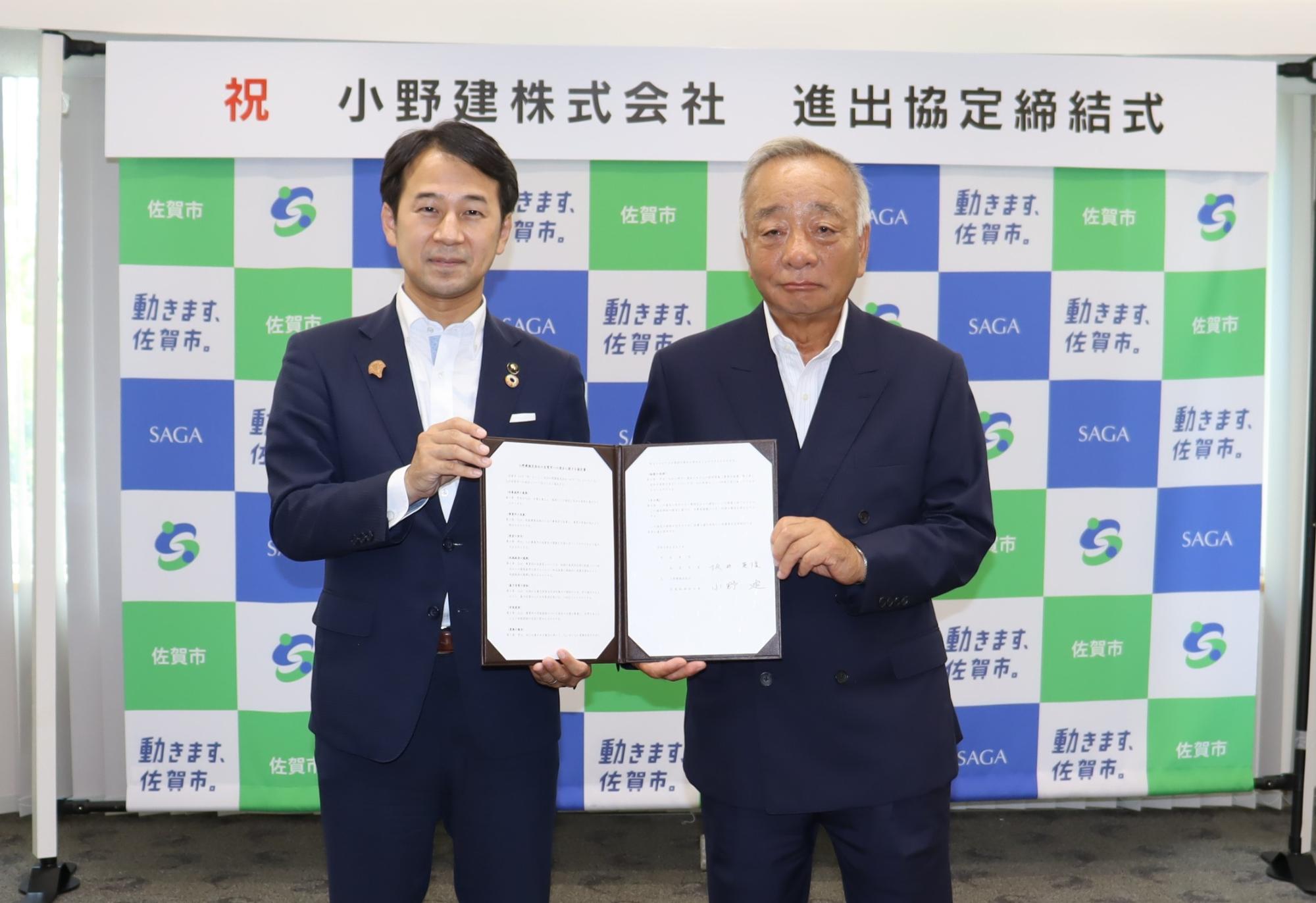 「祝 小野建株式会社 進出協定締結式」という看板を背に、佐賀市のロゴが格子状に並ぶ背景の前で、スーツ姿の市長と代表の男性が見開きになった協定書を一紙に持ち記念撮影をしている写真