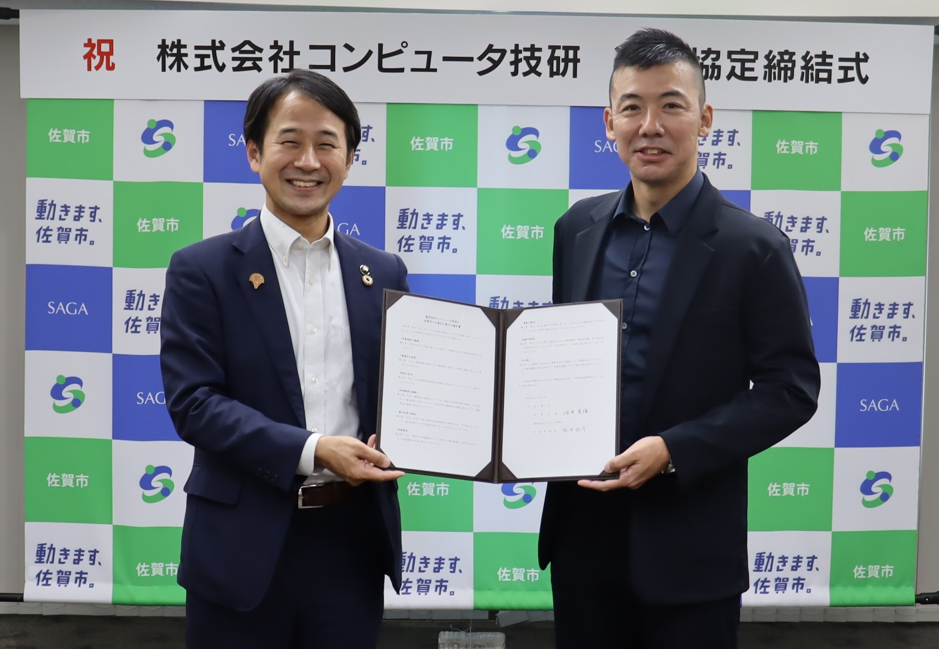「祝 株式会社コンピュータ技研 進出協定締結式」という看板の下、佐賀市のロゴが並ぶ背景の前で、スーツ姿の市長と代表の男性が見開きになった協定書を一緒に持ち記念撮影をしている写真