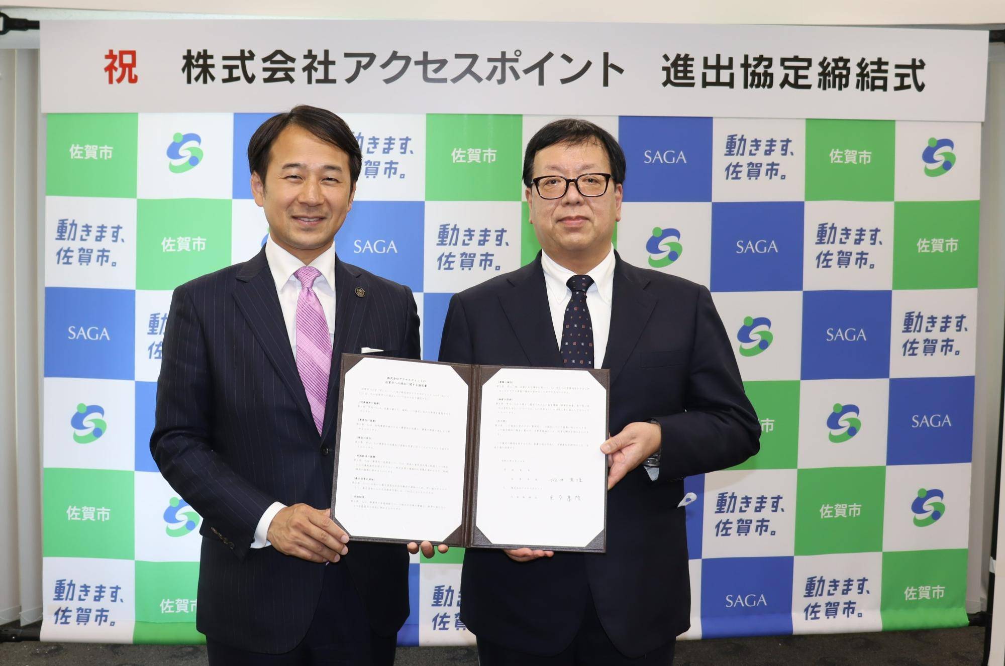 「祝 株式会社アクセスポイント 進出協定締結式」という看板を背に、佐賀市のロゴが格子状に並ぶ背景の前で、スーツ姿の市長と代表の男性が見開きになった協定書を一緒に持ち記念撮影をしている写真