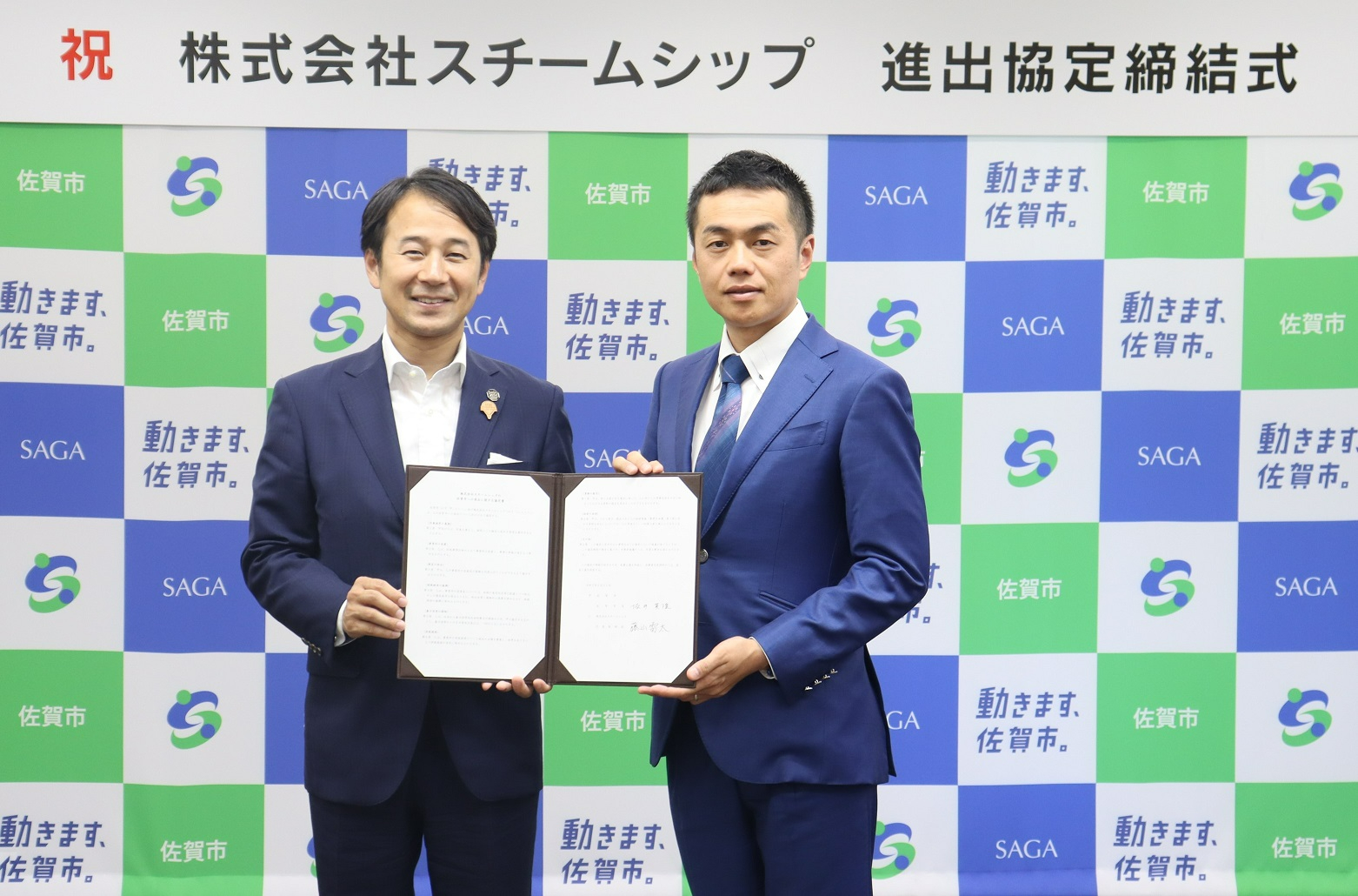 「祝 株式会社スチームシップ 進出協定締結式」という看板を背に、佐賀市のロゴが格子状に並ぶ背景の前で、スーツ姿の男性二人が見開きになった協定書を一緒に持ち記念撮影をしている写真