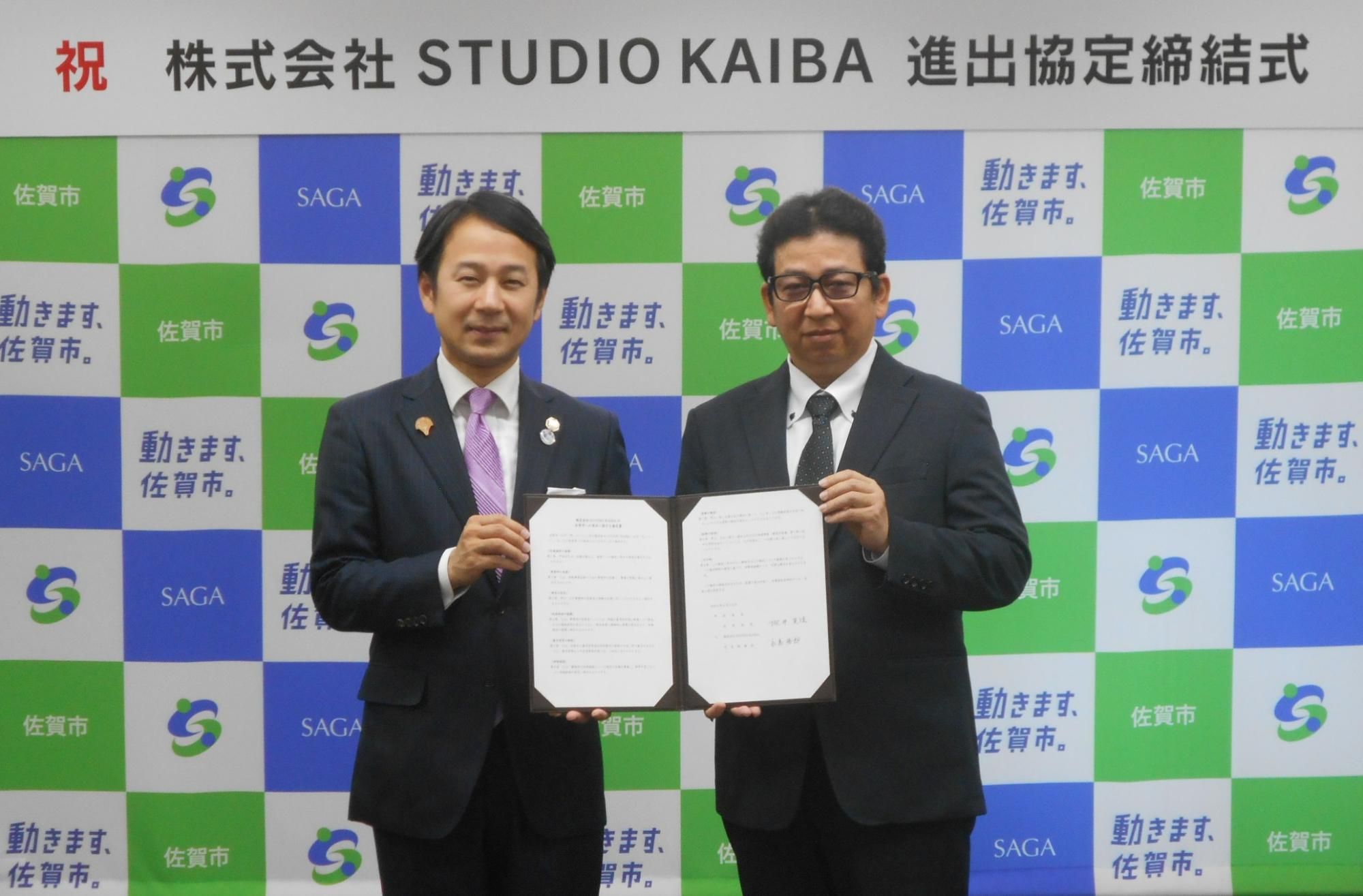 「祝 株式会社STUDIO KAIBA 進出協定締結式」という看板の下、佐賀市のロゴが格子状に並ぶ背景の前で、スーツ姿の男性二人が見開きになった協定書を一緒に持ち記念撮影をしている写真