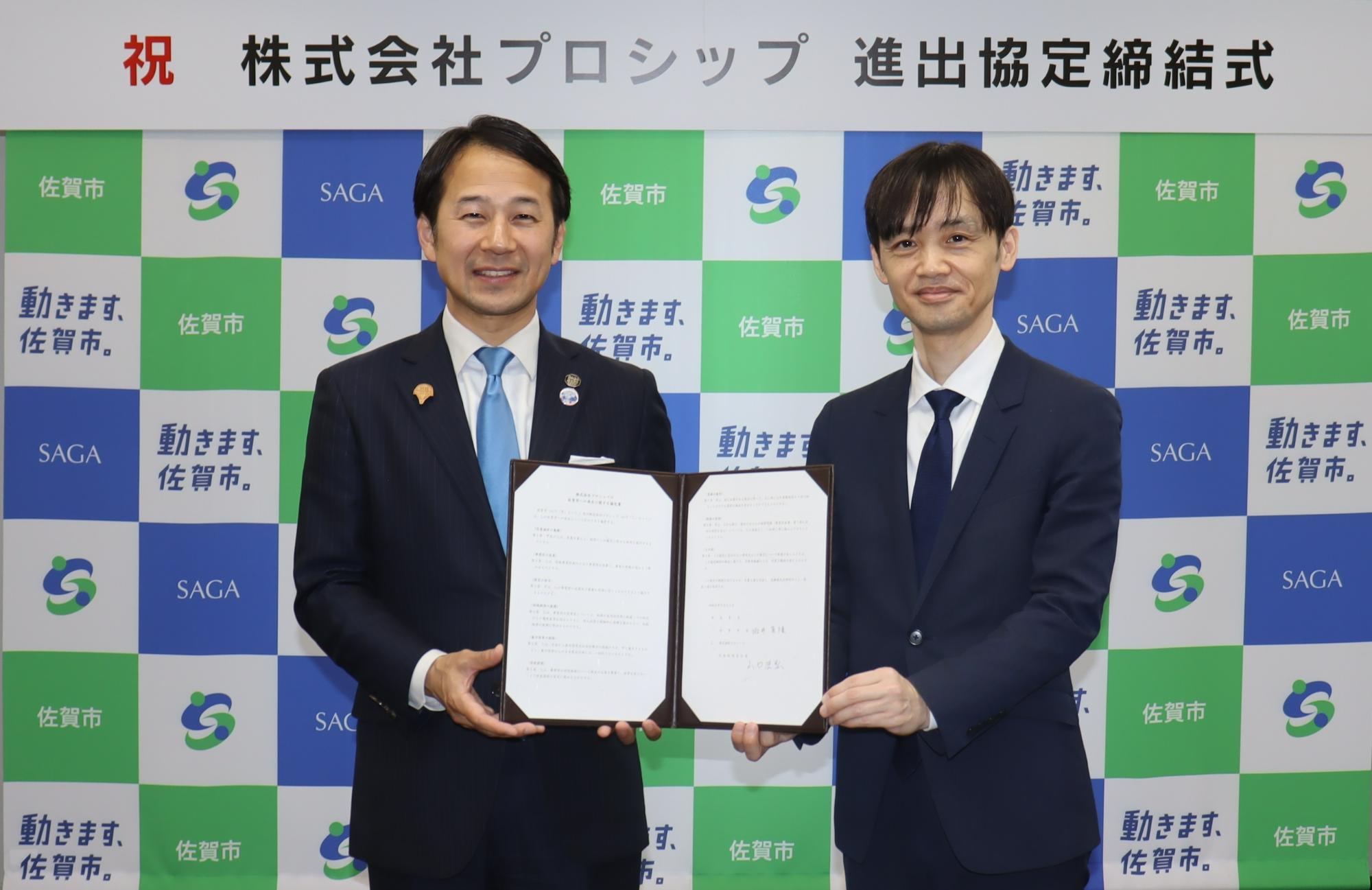 「祝 株式会社プロシップ 進出協定締結式」という看板を背に、佐賀市のロゴが格子状に並ぶ背景の前で、スーツ姿の男性二人が見開きになった協定書を一緒に持ち記念撮影をしている写真