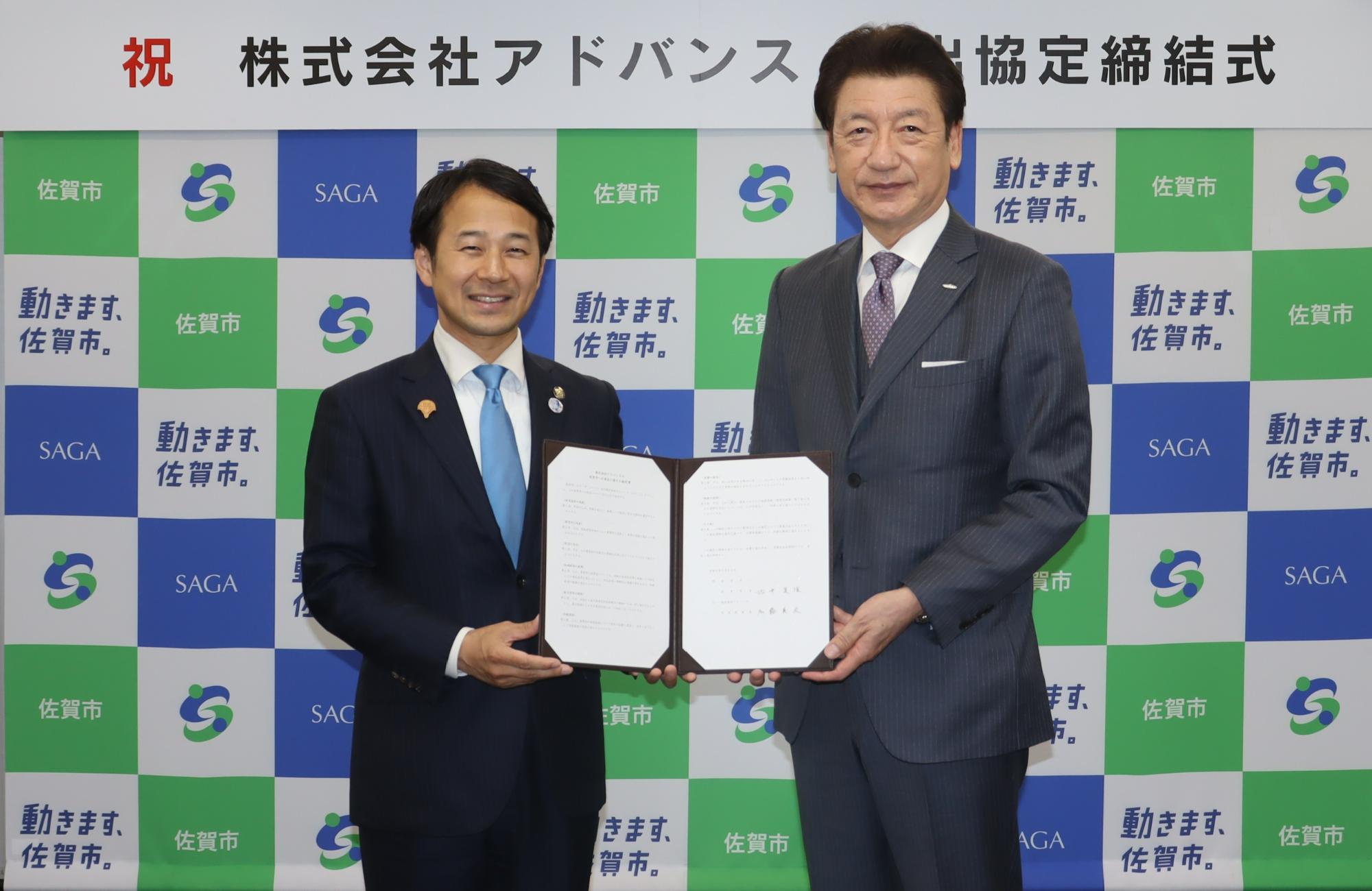 「祝 株式会社アドバンス 進出協定締結式」という看板の下、佐賀市のロゴが描かれた背景の前で、スーツ姿の男性二人が開いた協定書を一緒に持ち記念撮影をしている写真