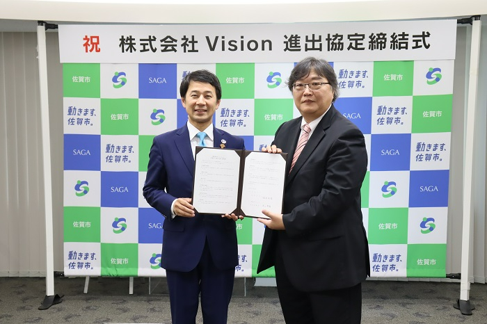 「祝 株式会社Vision 進出協定締結式」と書かれた横断幕を背に、佐賀市のロゴが並ぶ背景の前で、、スーツ姿の男性二人が協定書を一緒に持ち記念撮影をしている写真