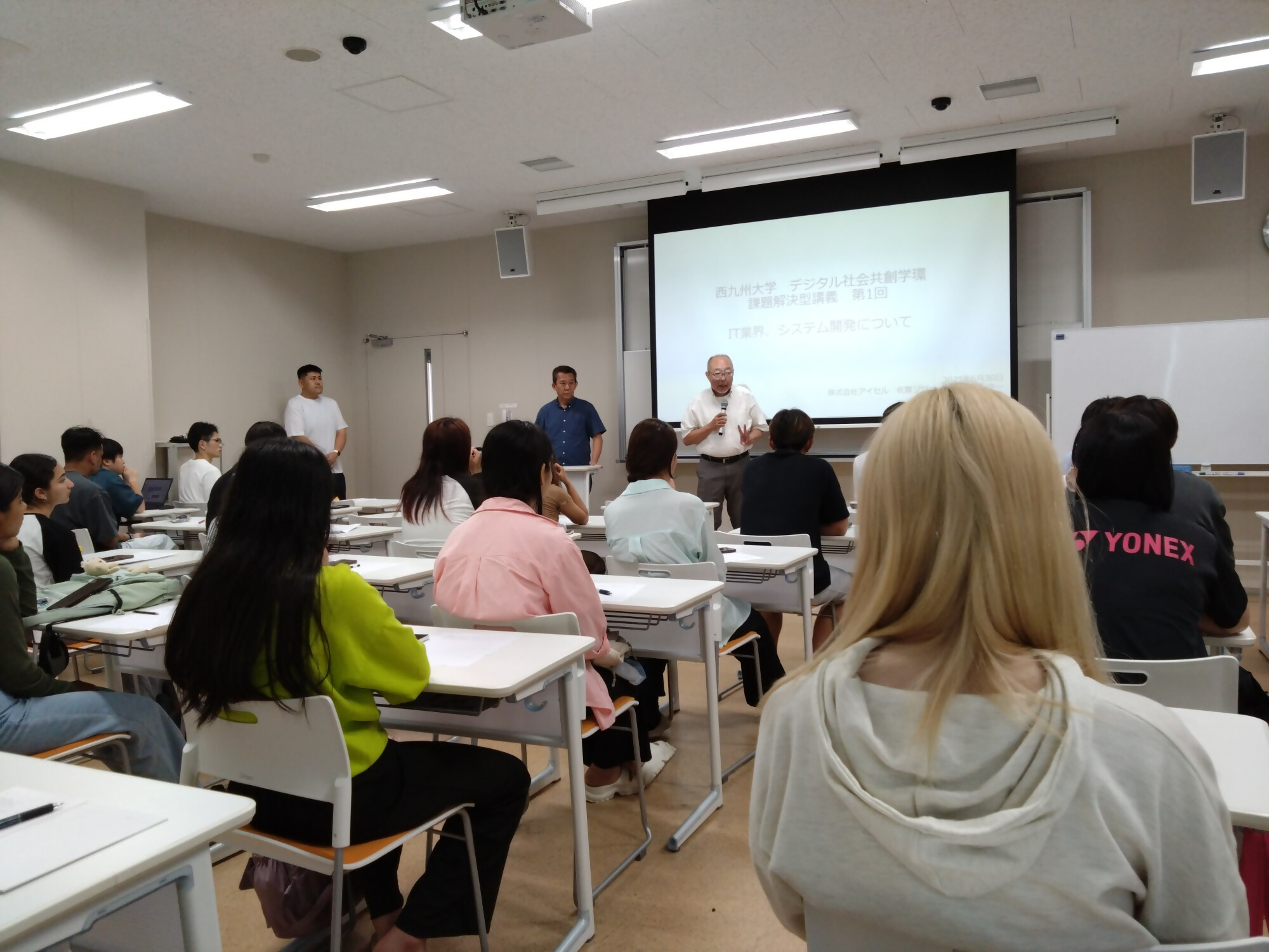 講師が「IT業界、システム開発について」というスライドを背にマイクで語りかけ、学生たちが机に向かって熱心に耳を傾けている様子の写真