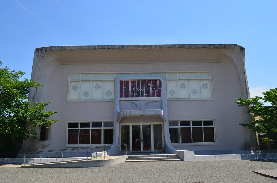 緩やかに曲線を描くコンクリートの大きな庇が特徴的な建物の正面外観で、中央の入り口上部には赤色の装飾窓があり、左右の窓には等間隔に家紋のような意匠が配置された、青空の下に佇むモダンな意匠の大隈重信記念館の建物外観の写真