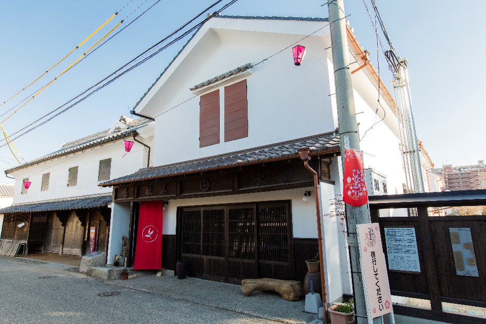 青空の下、白い漆喰壁が美しい二階建ての蔵造りの建物で、重厚な瓦屋根の庇や木製の格子戸が並び、軒先にはピンク色の提灯が吊るされた、歴史ある街道の情緒と華やかさが調和した「旧森永家」の風景写真