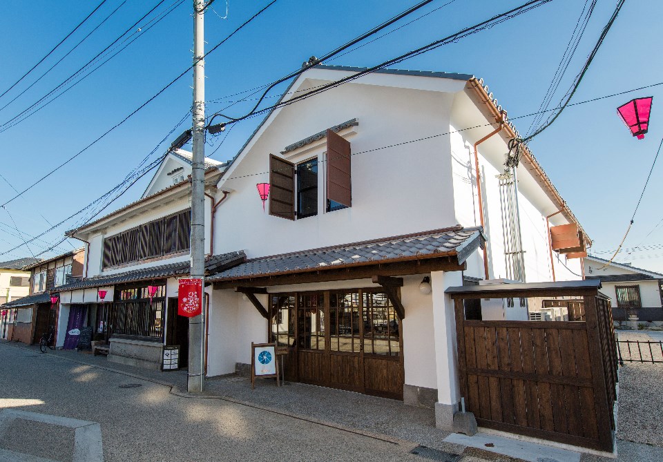 青空を背に建つ白壁の二階建て蔵造り建築で、瓦屋根の庇の下に木製の引き戸が並び、二階の開かれた窓やピンク色の提灯が街歩きの楽しさを演出している、歴史と現代が調和した「旧久富家」の風情ある建物写真