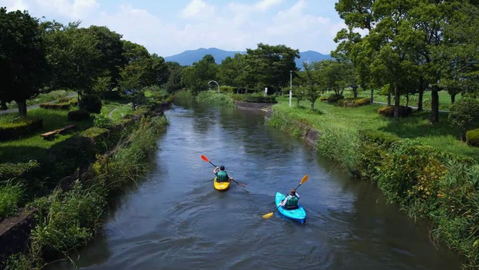 豊かな緑に囲まれた穏やかな川を、黄色と青色のカヤックに乗った二人の人物がオレンジ色のパドルを漕いで進んでおり、遠くの山並みと青空が調和した爽やかな夏のレジャーシーンを捉えた風景写真