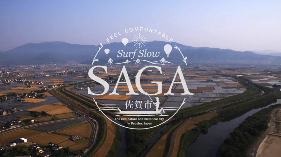 広大な佐賀平野の田園風景と山々を背景に、中央にバルーンや木々のイラストを添えた「SAGA 佐賀市 Surf Slow」などの文字が白抜きのエンブレム風にデザインされたロゴマーク