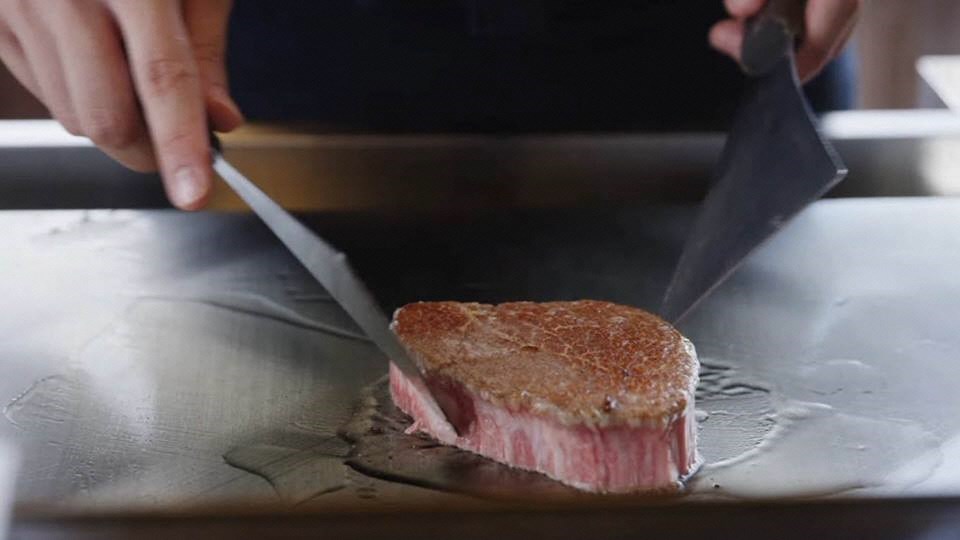 厚みのある霜降りの佐賀牛が鉄板の上でこんがりと焼かれ、料理人が両手に持ったナイフとヘラを使って手際よく肉の状態を確認しながら調理を進めている、食欲をそそる臨場感たっぷりの鉄板焼きの料理写真