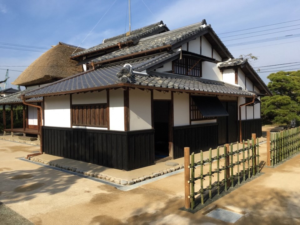 白壁と黒い腰壁のコントラストが美しい伝統的な日本家屋の建物で、手前には竹垣が並び、奥には瓦屋根の主屋と茅葺き屋根の一部が青空の下で見事に調和している様子を斜め前から捉えた、大隈重信生家の写真
