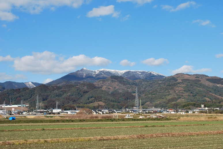 青空の下、冠雪した遠くの山並みを背景に、緩やかな斜面の山々とふもとに広がる集落、そして手前にまっすぐ区画された広大な田園風景が幾重にも重なり合う、自然豊かなのどかな風景写真