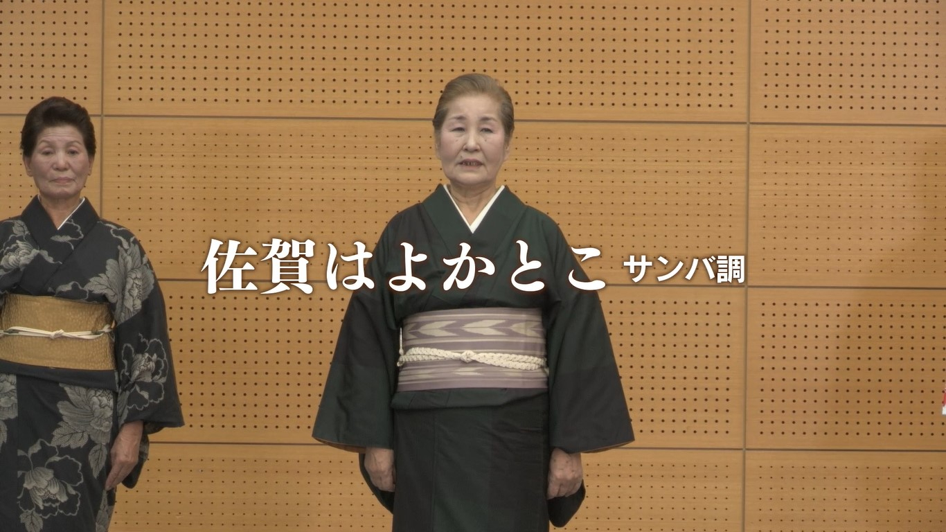 中央には大きく「佐賀はよかとこ サンバ調」という白い文字が重なるように配置され、格子模様の壁を背景に、黒い着物を凛々しく着こなした年配の女性が中央に立っている「佐賀はよかとこ」の踊り講習映像が表示された画面キャプチャ（佐賀城下栄の国まつり 佐賀はよかとこ（サンバ調） 講習ビデオ（YouTube））
