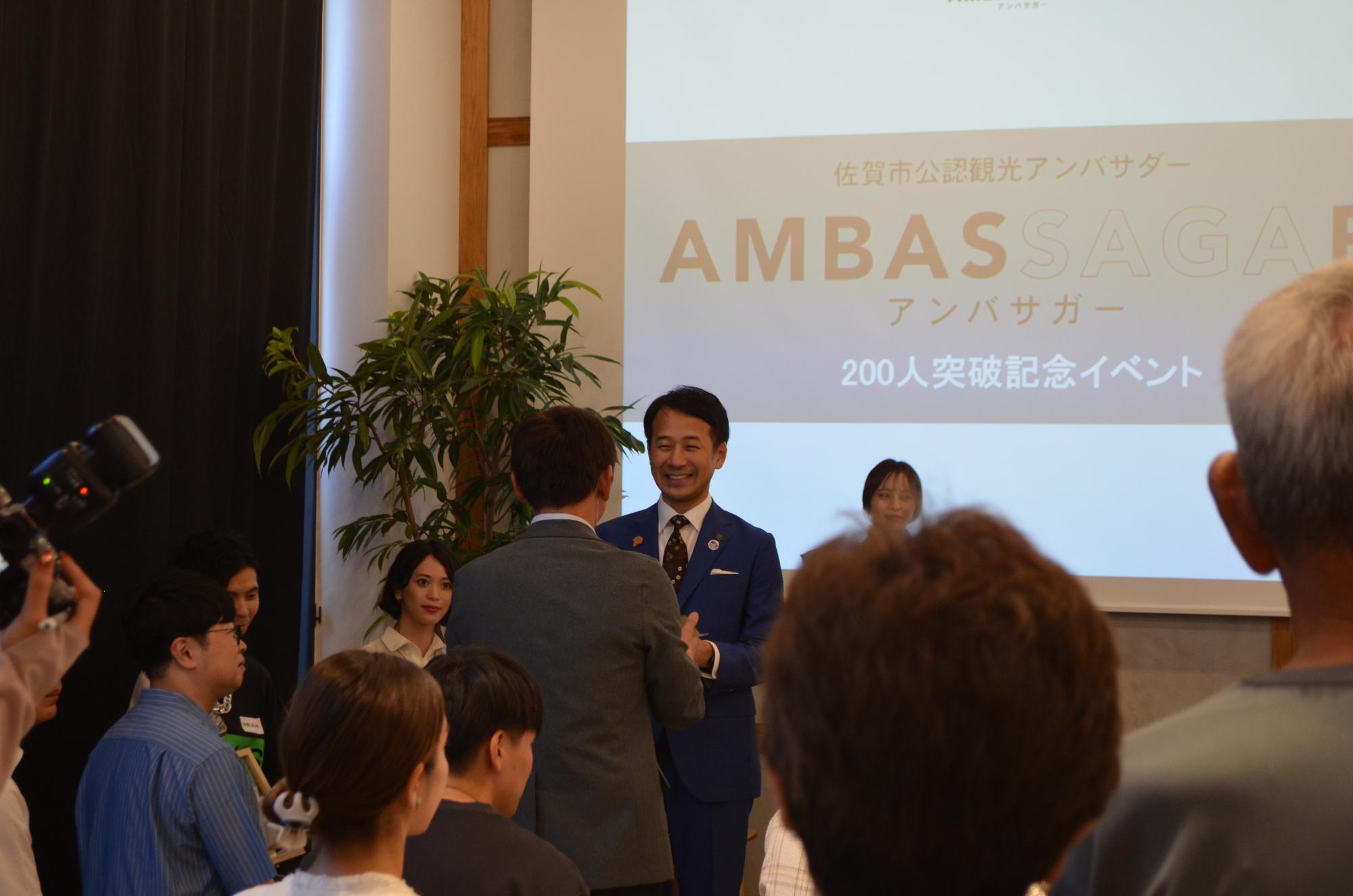 「佐賀市公認観光アンバサダー AMBASSAGAR アンバサダー 200人突破記念イベント」と記されたスクリーンを背景に、青いスーツ姿の市長が笑顔で参加者と交流する、会場の熱気と達成感を捉えたイベントの記録写真
