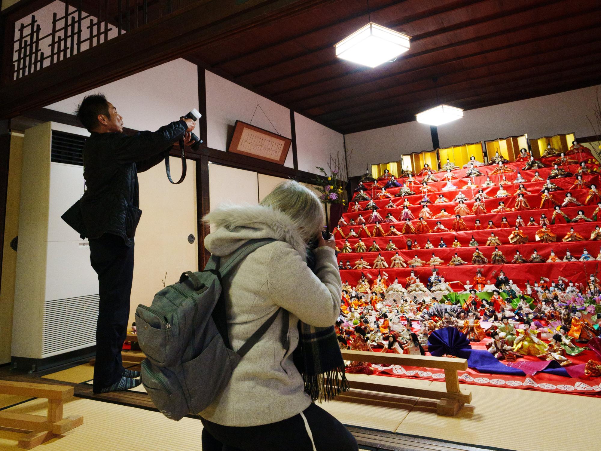 薄暗い和室の奥に飾られた、豪華な金屏風を背景に何十体もの雛人形が並ぶ巨大な七段飾りの雛壇に向かって、手前の畳の上で一眼レフカメラを構えて熱心に撮影を続ける男女の姿を背後から捉えた撮影の様子の写真