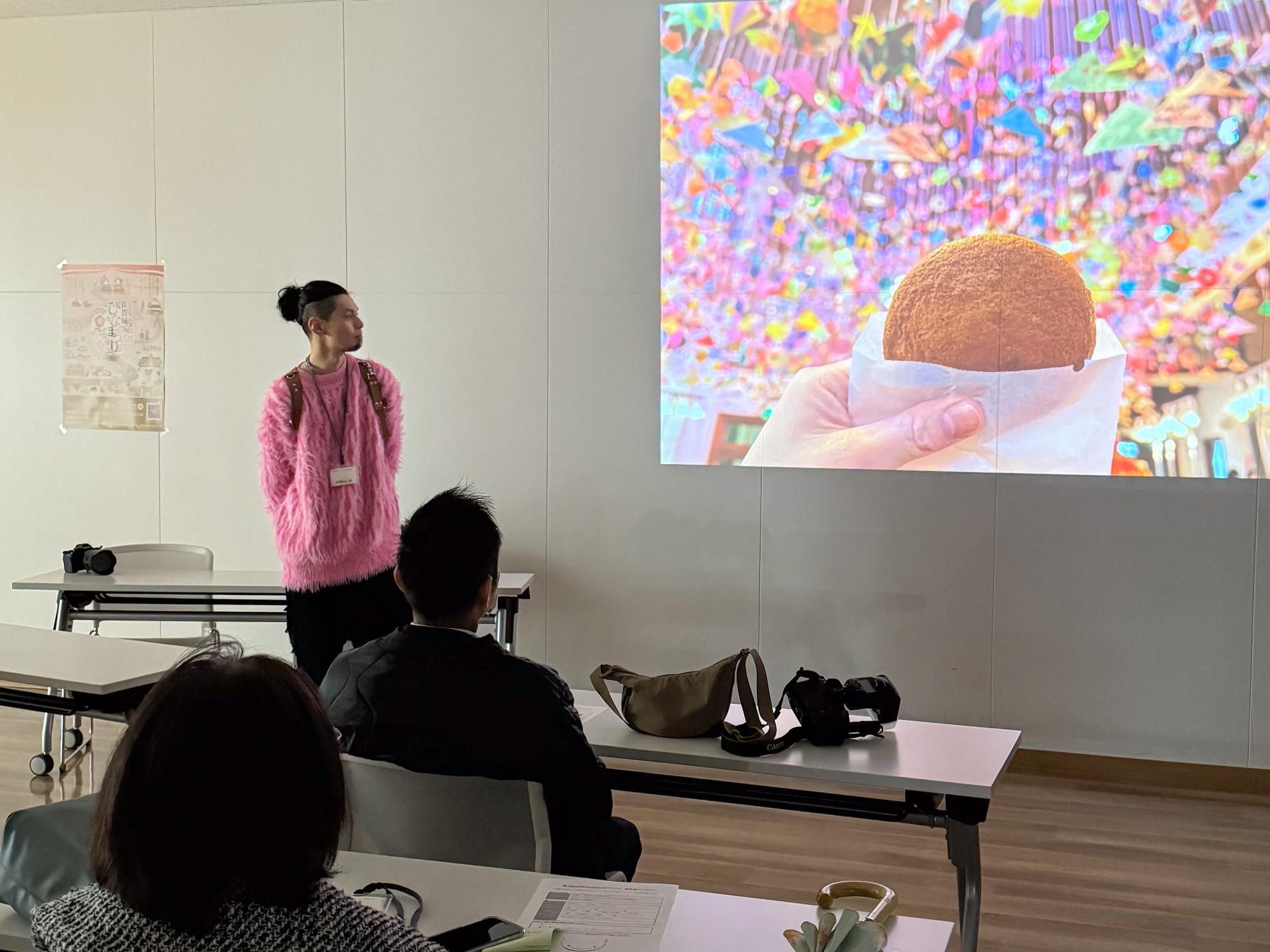プロジェクタースクリーンに映し出された色鮮やかな吊るし雛と丸い菓子の写真を背に、鮮やかなピンク色のシャギーニットを身にまとった男性が立ち、手前の座席から参加者がその様子を見守っている、講評会のひとコマを捉えた写真