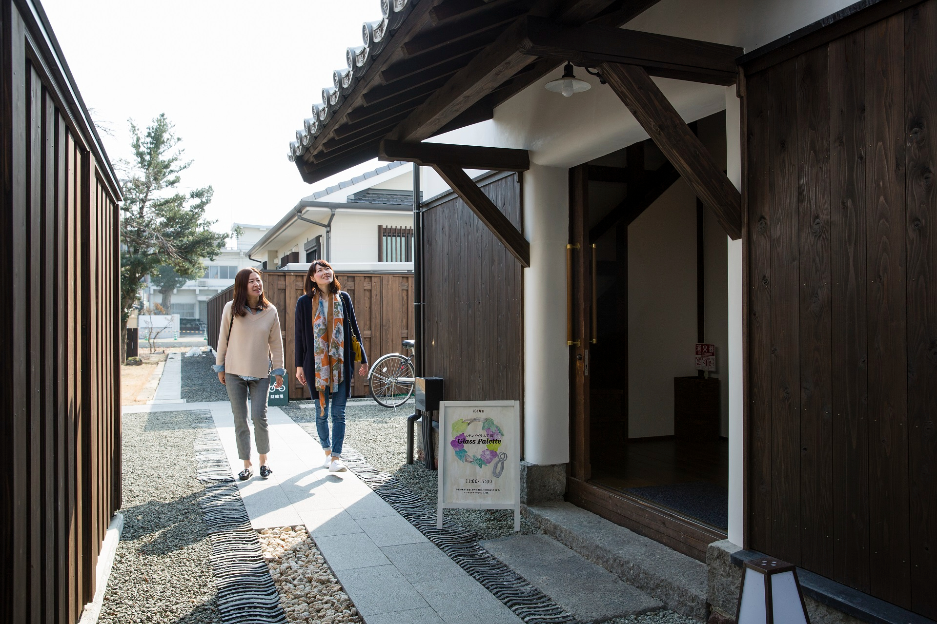 趣ある蔵造りの建物を背景に、カジュアルな装いの二人の女性が、石畳の小道を歩きながら伝統的な街並みの散策を和やかに楽しんでいる、旧久富家通路を捉えた写真