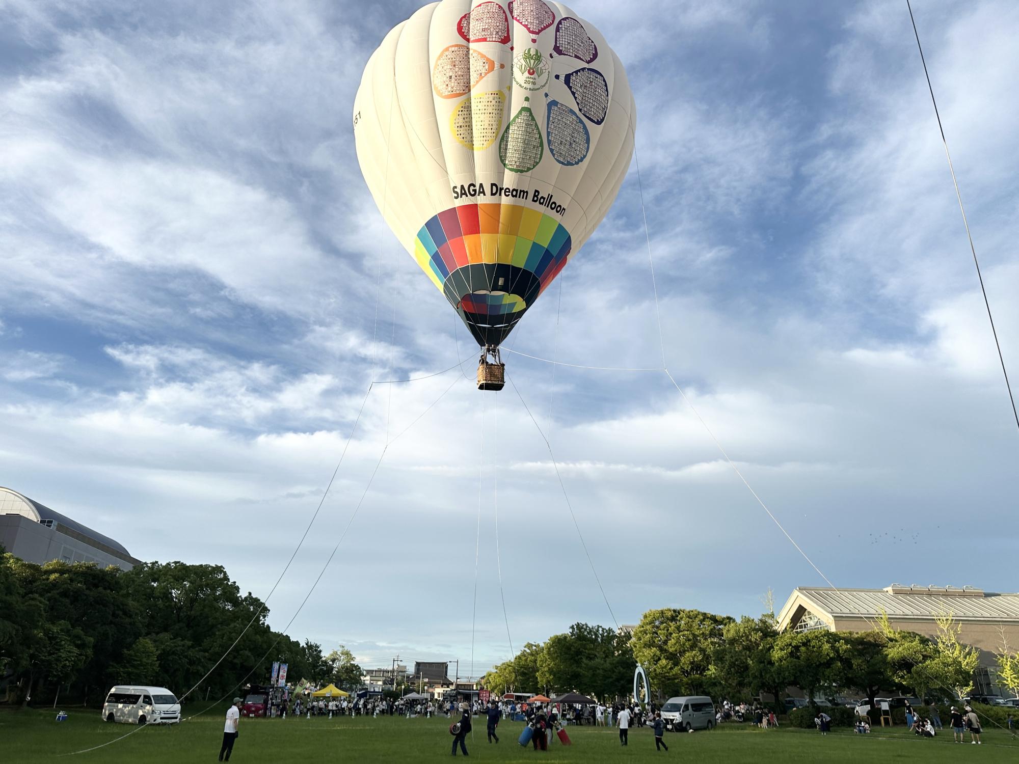 芝生が広がる会場で虹色の装飾と中央に花のようなロゴ、「SAGA Dream Balloon」の文字が描かれた大きな熱気球が、数本のロープで地上に繋がれて浮かんでいる係留飛行の様子を捉えた写真