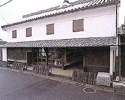 白い漆喰壁が美しい2階建ての伝統的な商家建築で、1階部分には格子戸や陳列棚が見える開放的な店構えがあり、2階には小さな窓が並び、重厚な瓦屋根と軒下の木造構造が歴史ある街並みの風情を今に伝える旧牛島家の写真