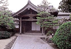 瓦屋根の伝統的な和風建築の玄関先を正面から捉えており、石畳の通路が建物へと続き、左右には丁寧に手入れされた松の木や低木が配置された趣のある日本庭園が広がっている、歴史的な風格を感じさせる旧古賀家の写真