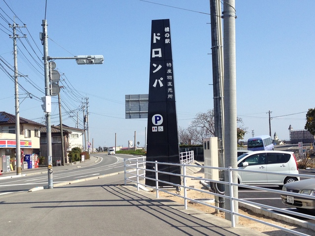 青空の下、道路沿いに立つ「橋の駅ドロンパ 特産物直売所」と書かれた黒く細長い自立看板を中心に、駐車場マークやトイレのピクトグラム、周囲の街並みや走行する車などを捉えた、施設の入り口を示す屋外の景観写真
