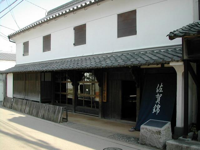瓦屋根の庇と白い漆喰壁が特徴的な二階建ての伝統的な商家建築で、入り口には「佐賀錦」と白文字で記された大きな紺色の暖簾が掛けられており、歴史ある街道の面影を今に伝える落ち着いた佇まいの旧牛島家の建物写真