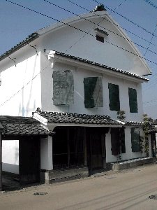 青空の下に広がる白い漆喰壁が美しい二階建ての蔵造りの建物で、二階部分には重厚な緑色の観音開きの扉が並び、一階の瓦屋根や伝統的な格子戸が歴史ある商家の面影を感じさせる、端正で凛とした佇まいの旧三省銀行の建築写真