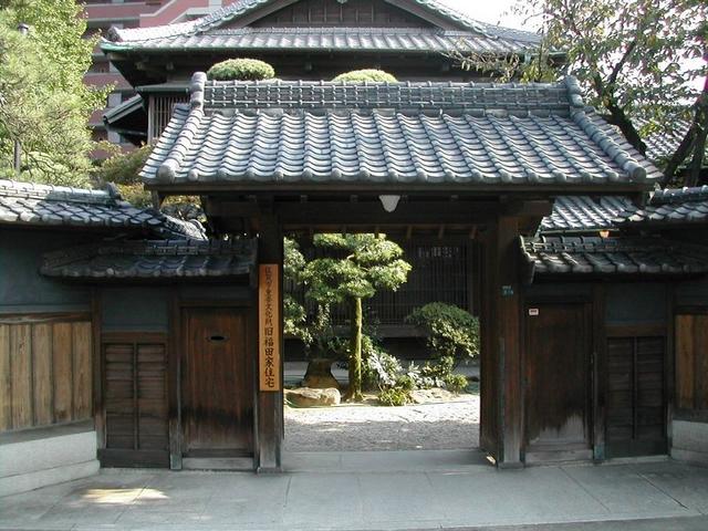 瓦屋根を冠した重厚な木造の門の奥に、美しく手入れされた松の木が立つ中庭と伝統的な日本家屋の主屋が広がり、歴史の重みと静謐な情緒を感じさせる「旧福田家住宅」の入り口を正面から捉えた風景写真