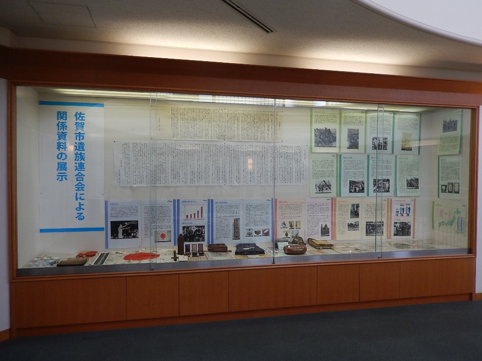 ガラス張りの大きな展示ケース内に「佐賀市遺族連合会による関係資料の展示」という文字が掲げられ、壁面には戦地別の状況や統計グラフ、底部には日章旗や軍服、千人針などの貴重な実物資料が所狭しと並べられた展示風景の写真