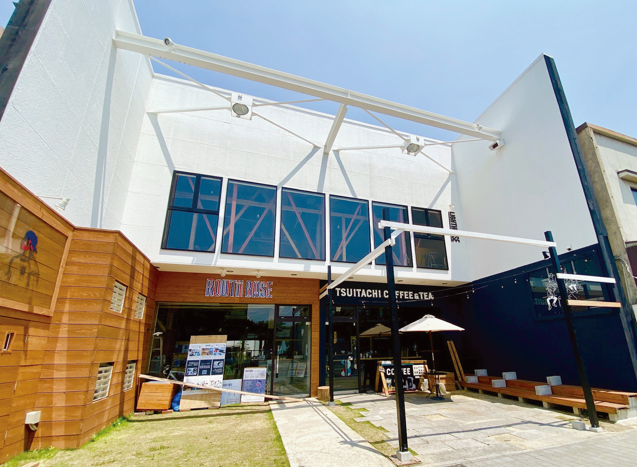 白い外壁と大きな窓を備えた建物前に木調の店舗入口やテラス席が配置され明るく開放的な雰囲気が広がるtojin242の建物外観の写真