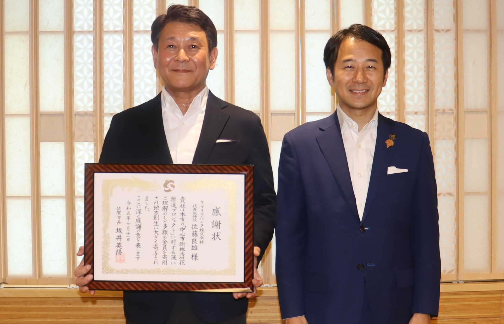 木格子の室内で男性二人が並び立ち、左の人物が丁寧に額装された感謝状を胸の前でしっかりと掲げて示し、落ち着いた雰囲気の中で授与の様子を伝える写真