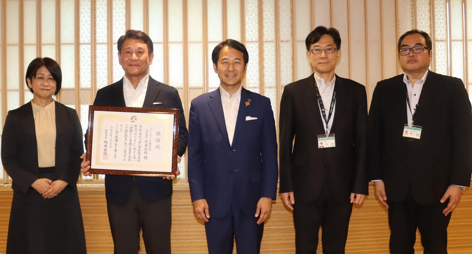 和風の木格子が並ぶ室内で男女五人が横一列に立ち中央の人物が額装の感謝状を両手で掲げ授与の雰囲気を伝える写真