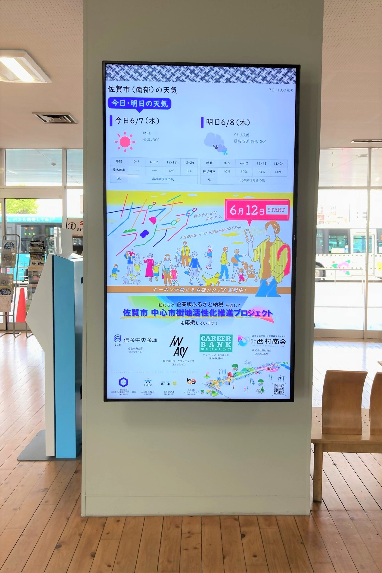屋内のデジタルサイネージに天気予報や日付情報とカラフルなイベント告知が大きく表示されている写真