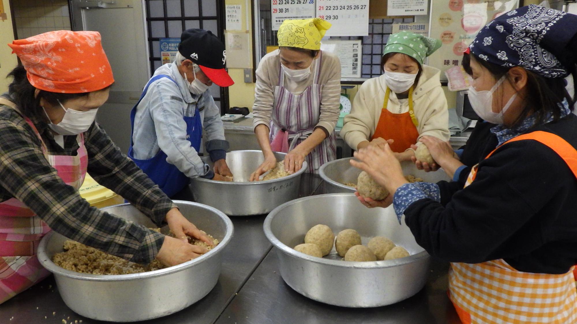 調理室で、お揃いの白い三角巾とマスクを着用した5人の女性たちが、大きな金属製のボウルを囲みながら談笑し、手際よく丸い生地のようなものを形成してボウルの中に並べている、活動的な風景の写真