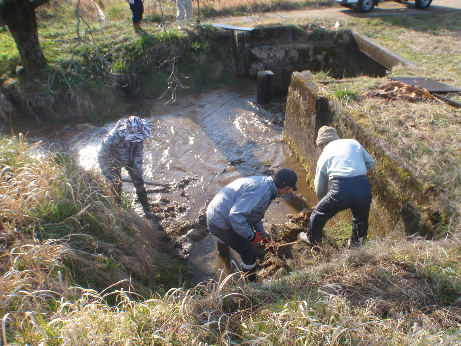 屋外の川や水路のような場所で、帽子を被った3人の作業員が泥の中に立ちながら、スコップや道具を使って堆積した土砂や泥を掻き出し、清掃や補修作業を行っている様子の写真