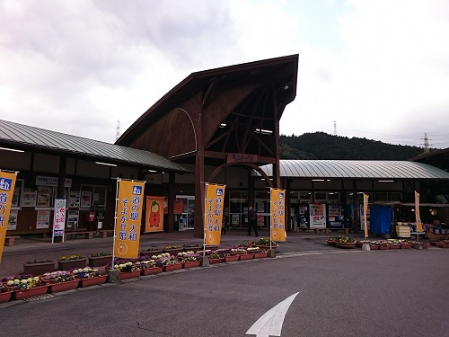 鋭角に尖った特徴的な木造屋根を持つ「道の駅 大和 そよかぜ館」の建物があり、入り口付近には黄色い「道の駅」ののぼり旗や色鮮やかな花が植えられた多くのプランターが並ぶ施設の外観を写した写真