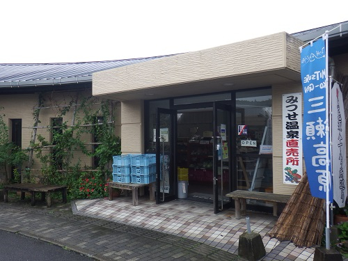 ベージュ色の外壁を持つ建物の入り口に「みつせ温泉直売所」という看板が掲げられ、青い「三瀬高原」ののぼり旗や積み上げられた水色の収穫カゴ、木製のベンチが置かれた、落ち着いた雰囲気の直売所の外観を写した写真