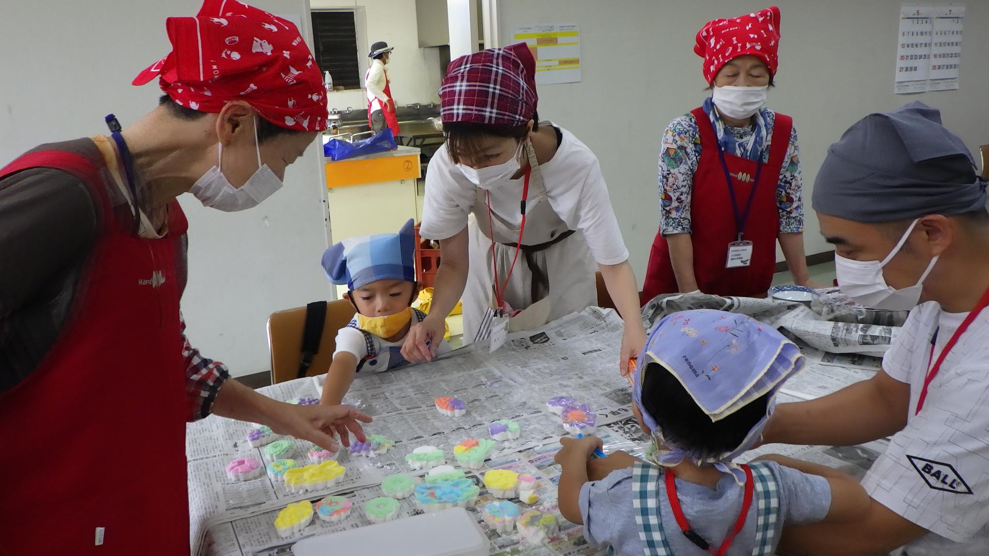新聞紙が敷かれた大きなテーブルを囲み、赤いエプロンとバンダナを身に着けた大人たちに見守られながら、小さな子供たちが色とりどりの粘土や型を使って熱心に工作を楽しんでいる屋内体験教室の様子を捉えた写真