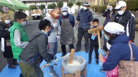 屋外の広場で青いビニールシートを敷き、中央に置かれた石臼を囲んで、マスク姿の大人や子供たちが交代で大きな木槌を振り下ろして餅つきを楽しみ、周囲で多くの人々がその様子を温かく見守っている行事風景を写した写真
