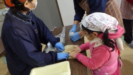 屋内の調理台の上で、紺色の上着を着た大人とピンク色の服に白いバンダナを巻いた小さな子供が、青い衛生手袋をはめて一緒にビニール袋に入った塊を丁寧に丸めている体験の様子を間近で捉えた写真