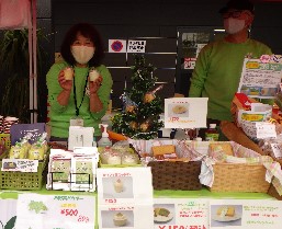 明るいグリーンの服を着てマスクをした女性が、小さなカップに入ったスイーツを両手に持って笑顔で披露しており、手前のテーブルにはシフォンケーキやプリンなどの焼き菓子が丁寧に並べられた、賑やかな催事販売の様子を写した写真