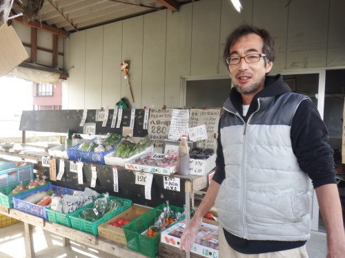軒下に並べられた木製の棚に、大根やブロッコリーなどの新鮮な野菜がカゴに入れられて並び、その横で眼鏡をかけた男性が穏やかな表情で立ち、手書きの値札や案内が掲げられた販売所を写した写真