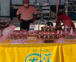 屋外のイベント会場で、黄色いテーブルクロスが掛けられた販売ブースの前に立ち、マスク姿の男性が腰に手を当てて接客しており、テーブルの上には瓶詰めのジャムや小分けにされた焼き菓子などがきれいに並べられている催事販売の様子を写した写真
