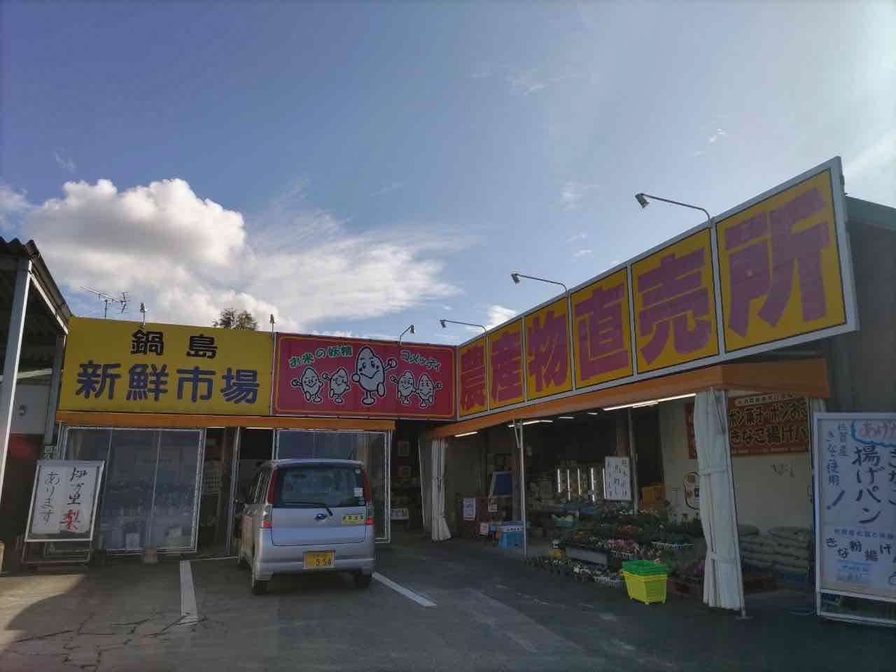 建物の屋根部分に「鍋島 新鮮市場」や「農産物直売所」と書かれた黄色い大きな看板が掲げられ、店頭には野菜の苗や商品が並び、駐車場にシルバーの軽自動車が停まっている活気ある直売所の外観を写した写真